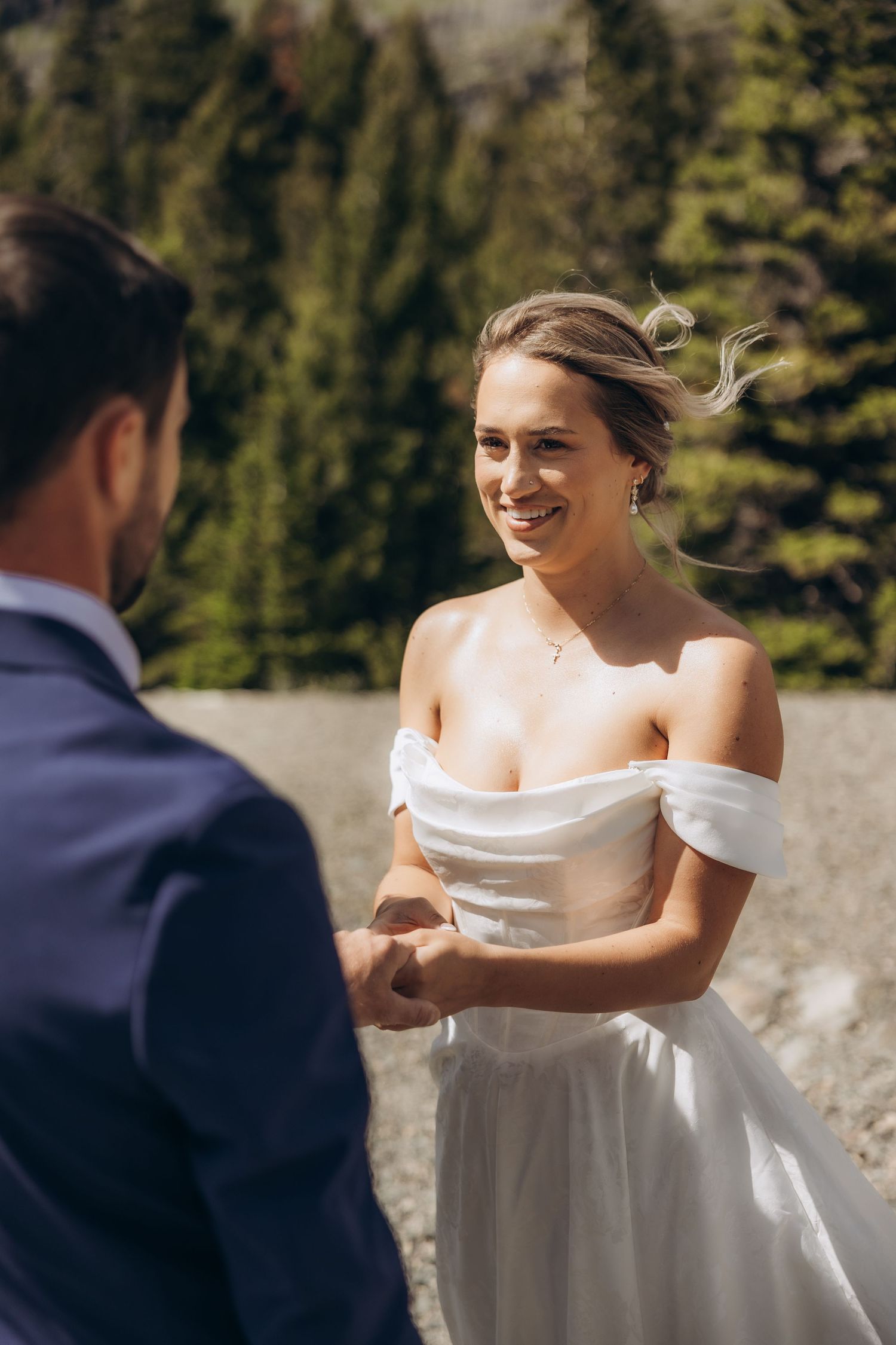 Smiling bride in off-shoulder white gown holds hands with groom in blue suit outdoors with pine trees.