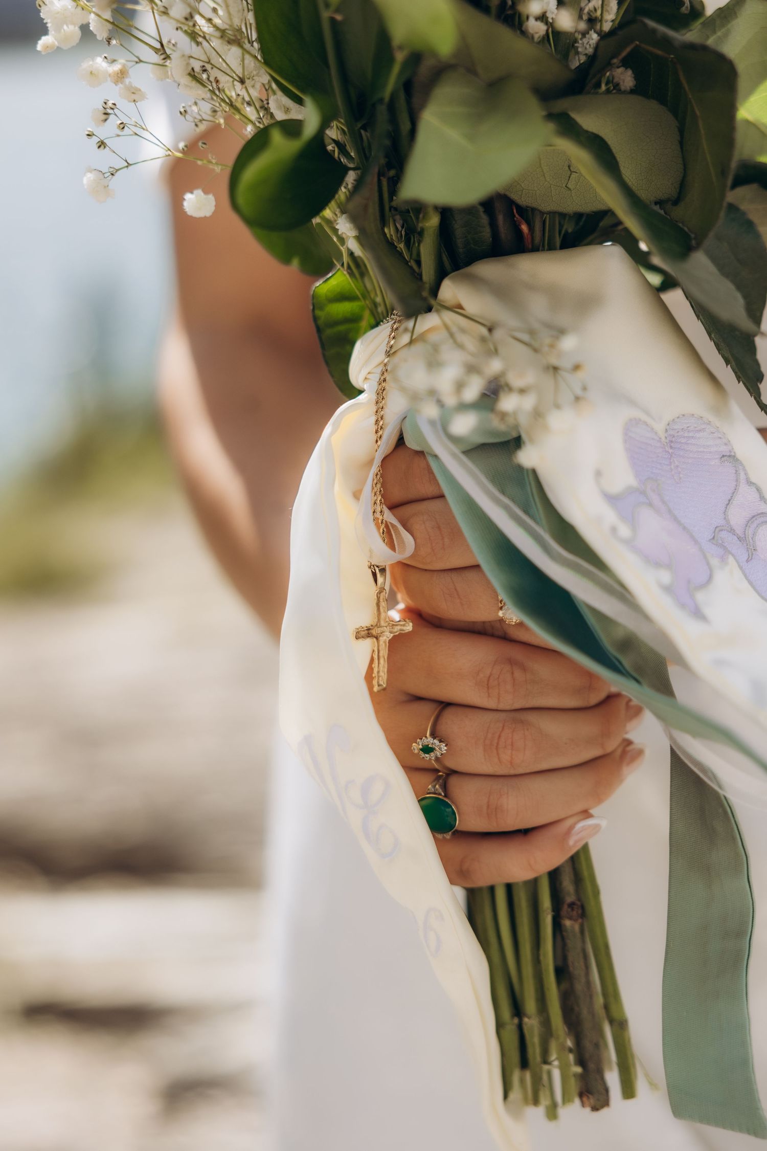 Bride holding a wedding bouquet of white flowers and greenery, adorned with gold rings and a cross charm.