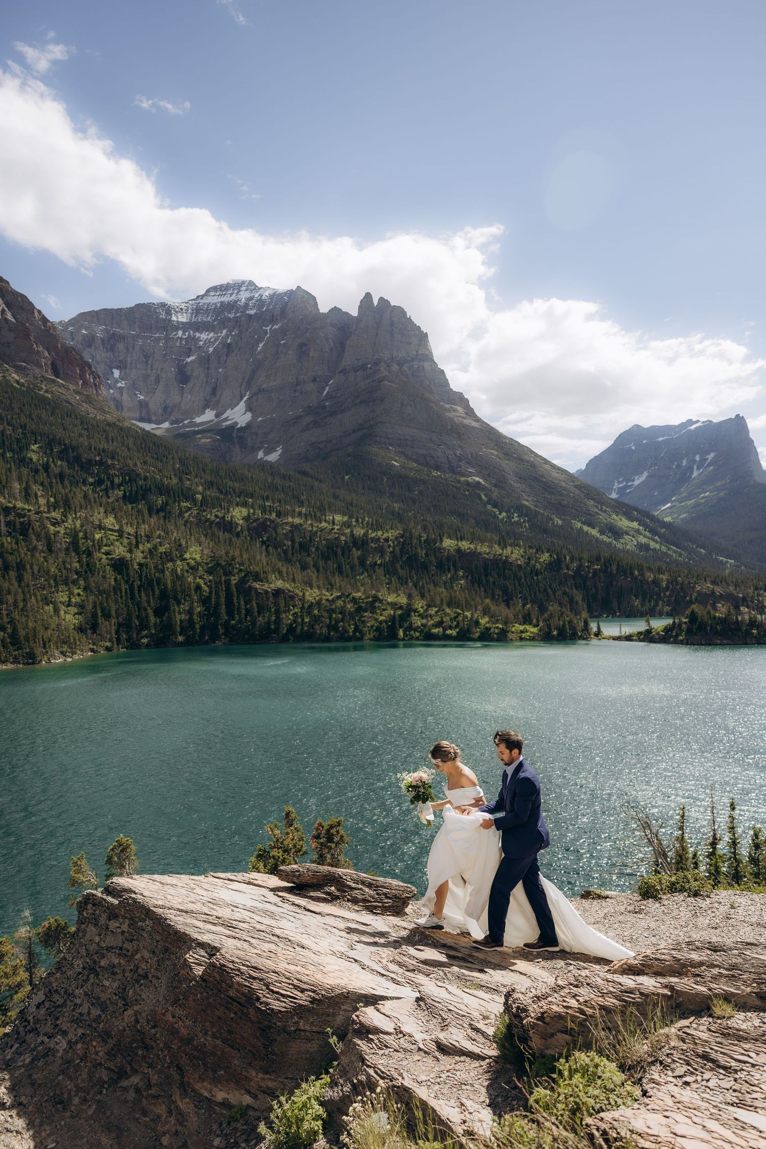 Bride and groom walk on rocky ledge overlooking turquoise mountain lake with peaks and forest backdrop.