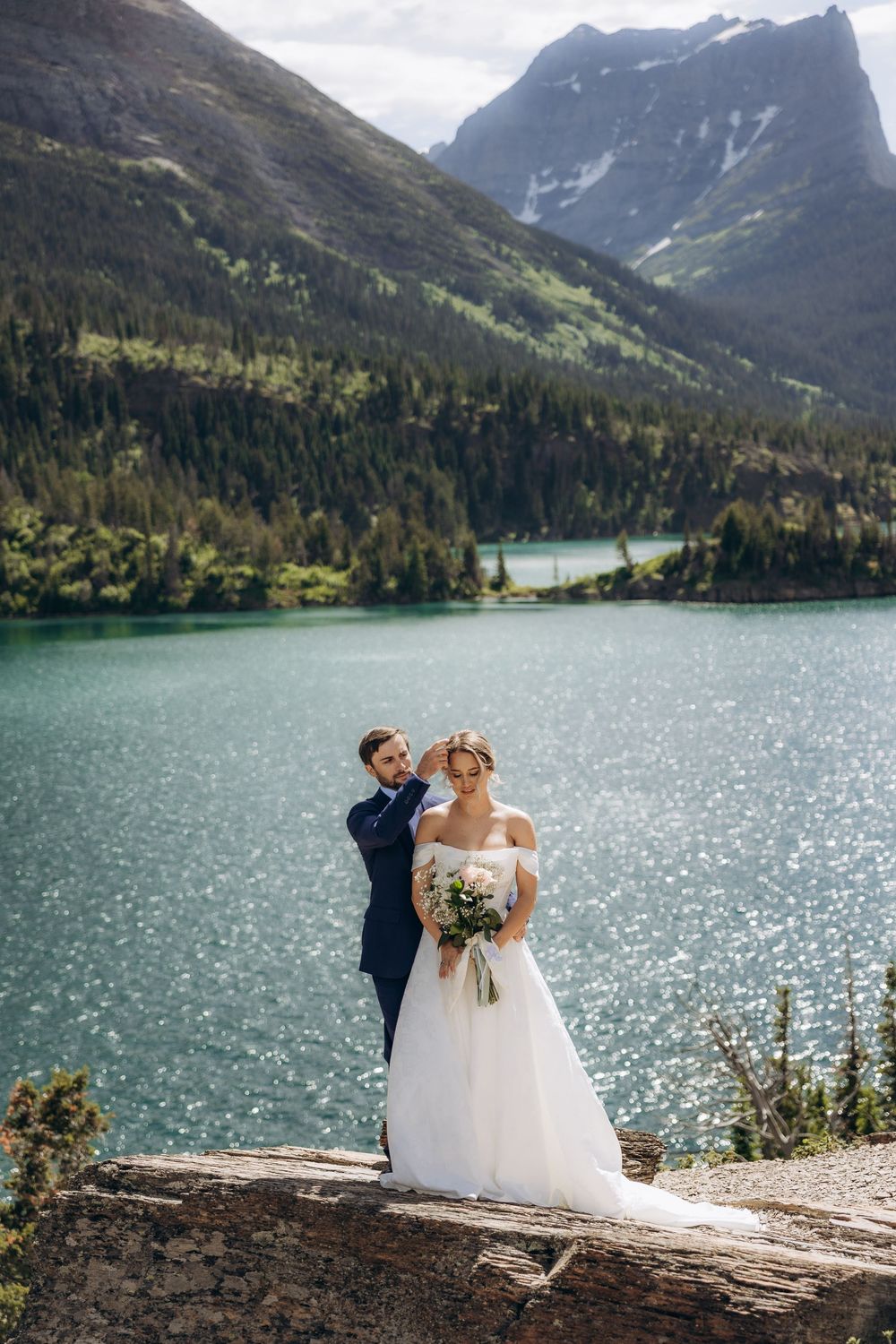 Bride and groom pose by a turquoise mountain lake, surrounded by pine forests and snow-capped peaks, bride holding a bouquet.