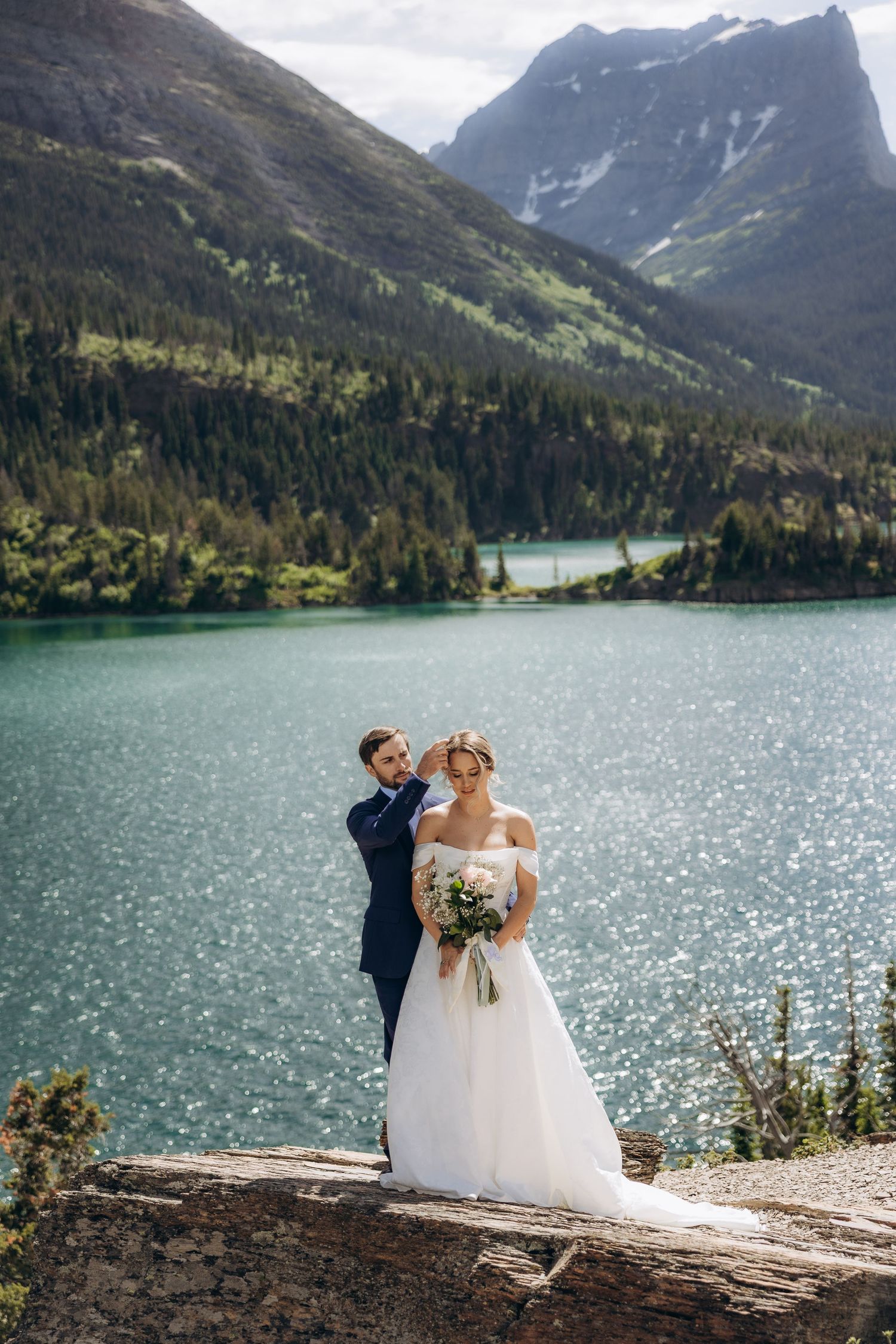 Bride and groom pose by a turquoise mountain lake, surrounded by pine forests and snow-capped peaks, bride holding a bouquet.