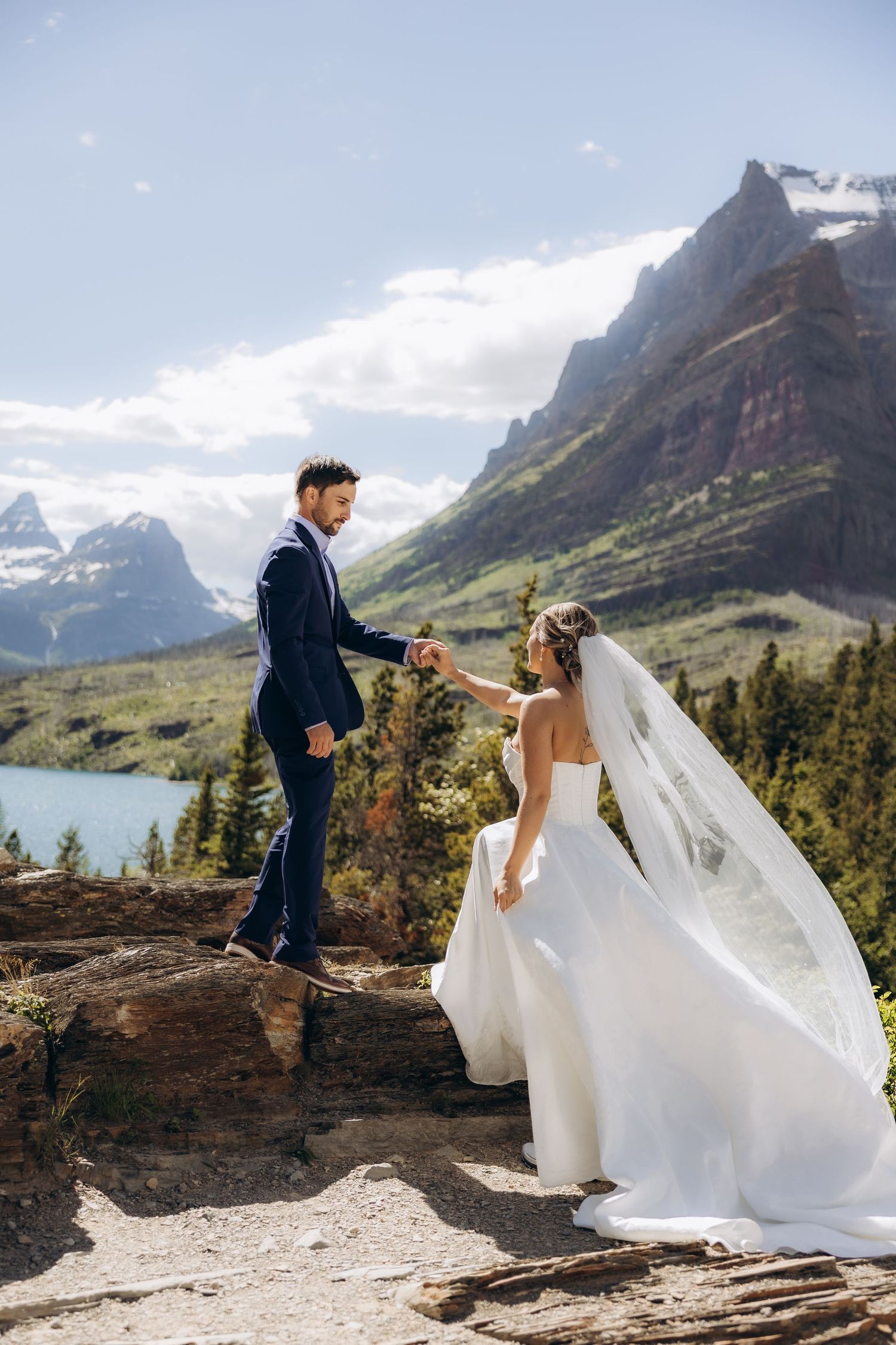 Bride and groom hold hands on rocky overlook with stunning mountain lake backdrop at Glacier National Park wedding.