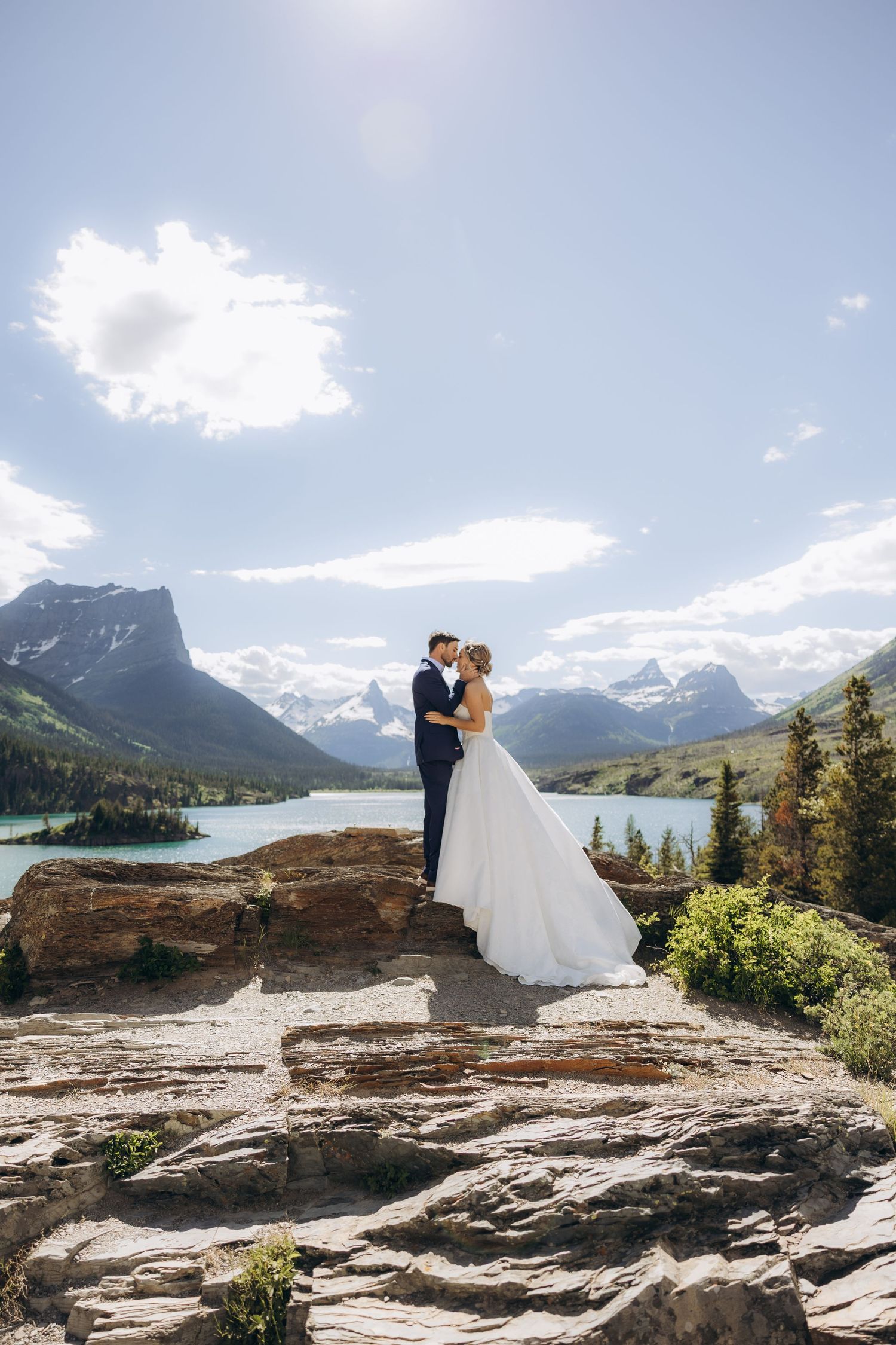 Bride and groom embrace on rocky mountain overlook with turquoise lake and snow-capped peaks backdrop on sunny day.