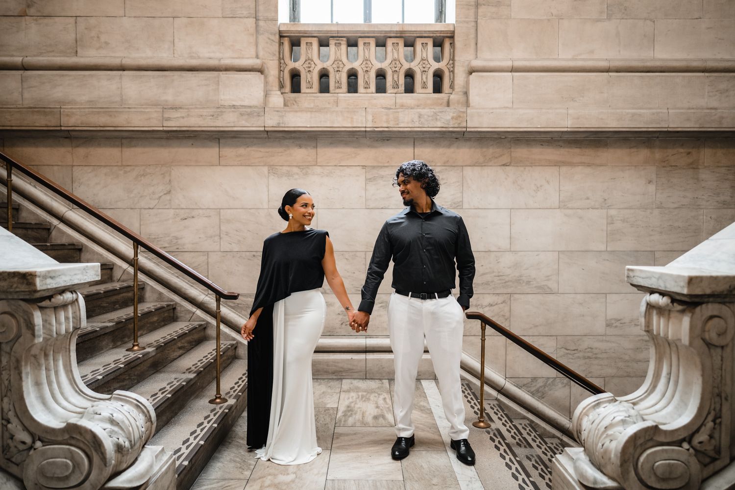 Elegant couple in black and white outfits holding hands on ornate marble staircase.
