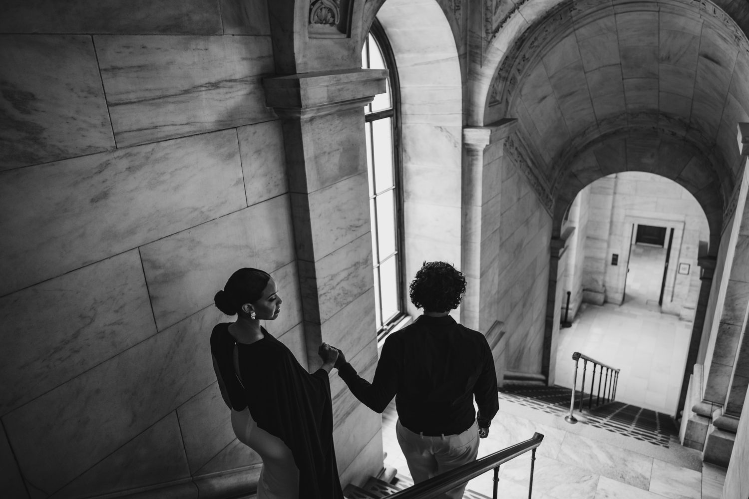 Black and white photo of a couple holding hands on grand marble staircase near arched window.