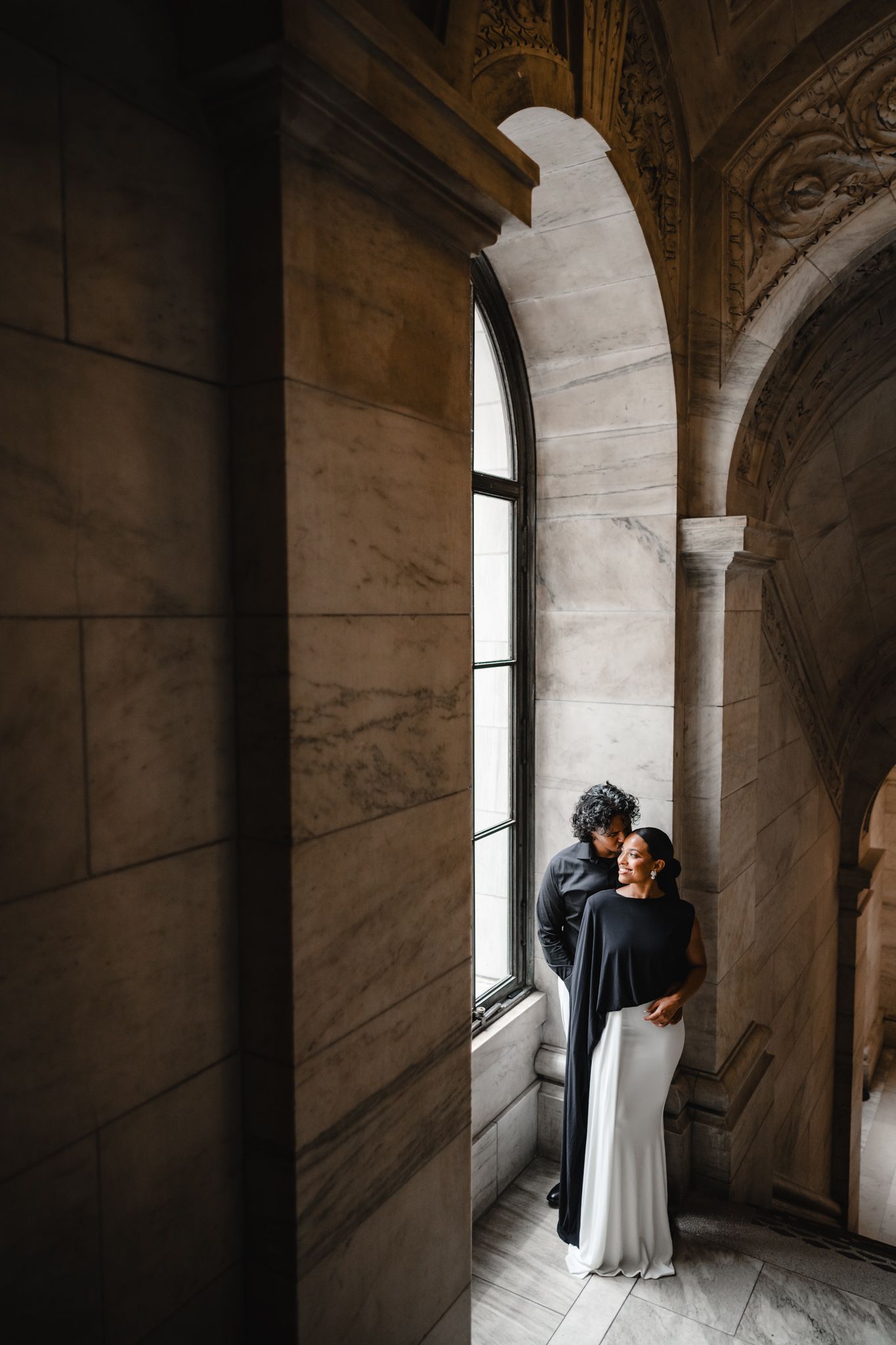 Couple embracing by an arched window in an ornate stone building, bride in white gown with black cape.