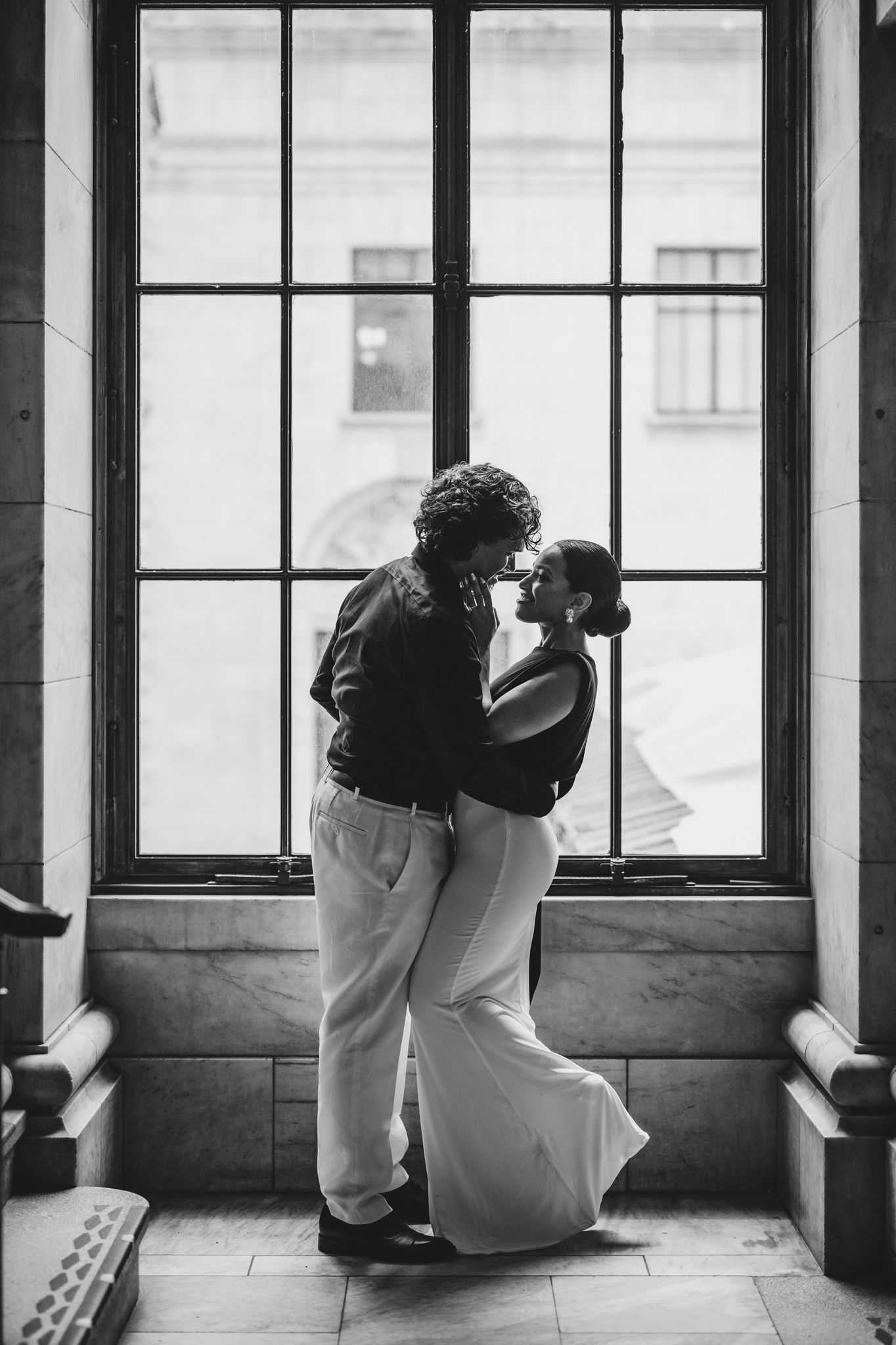 Black and white photo of a couple sharing a romantic moment, nearly kissing in front of a large window.
