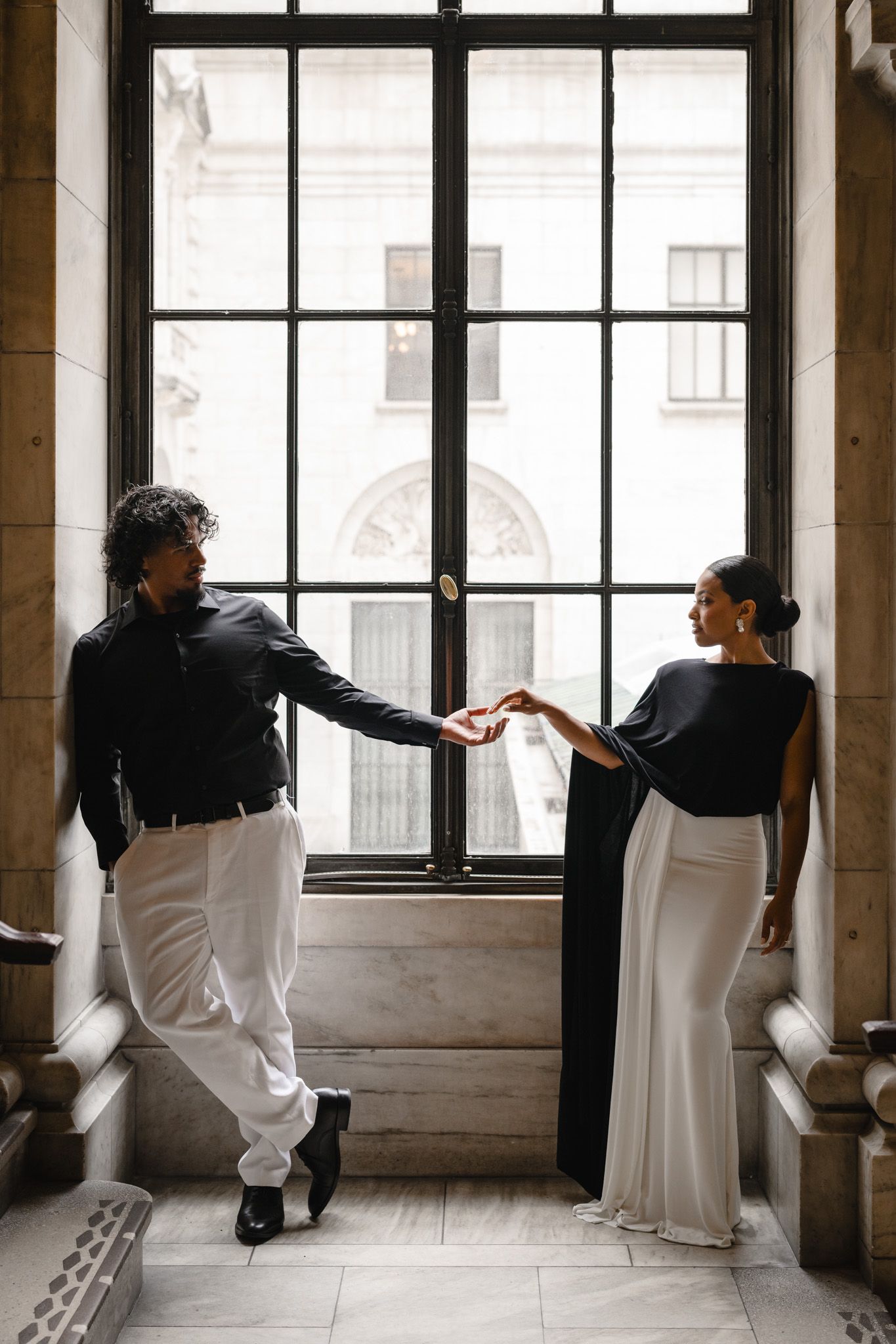 Elegant couple in black and white attire holding hands by a grand window in a classic architectural setting.