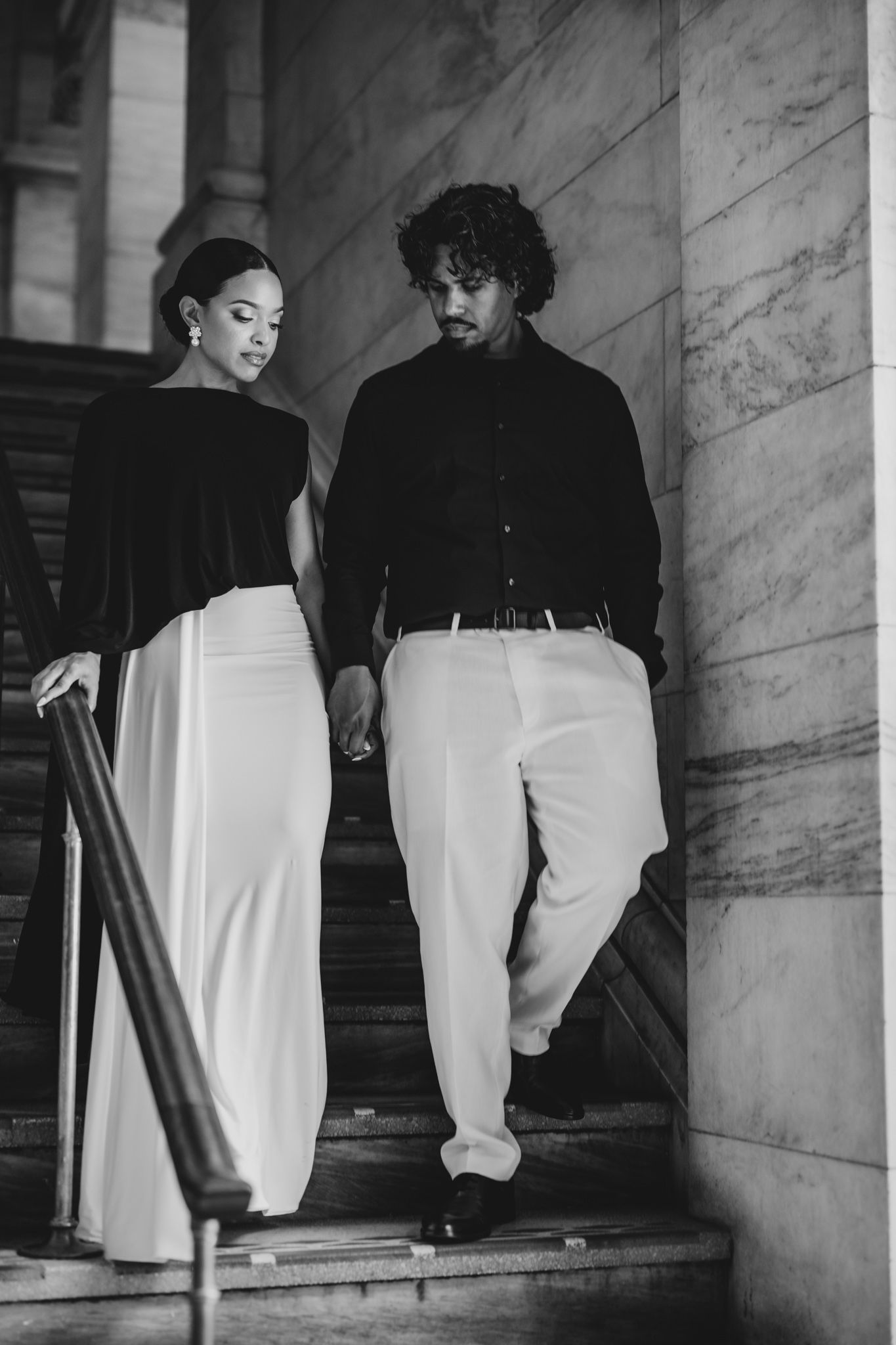 Elegant couple in black and white outfits descending marble stairs in a classic architectural setting.