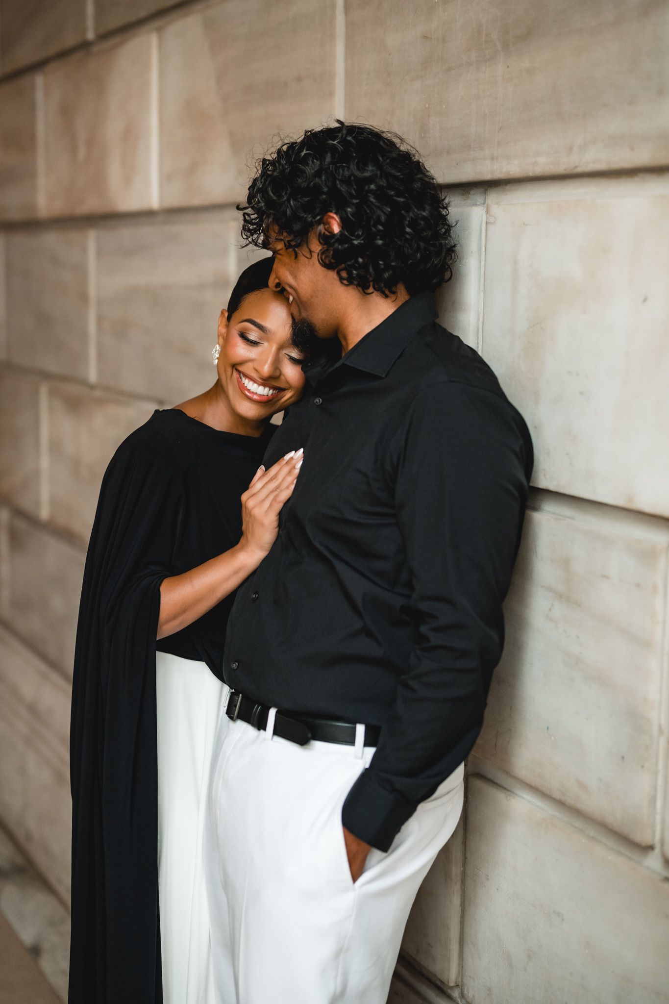 A smiling Black woman in a black dress leans into a man in a black shirt and white pants against a stone wall.