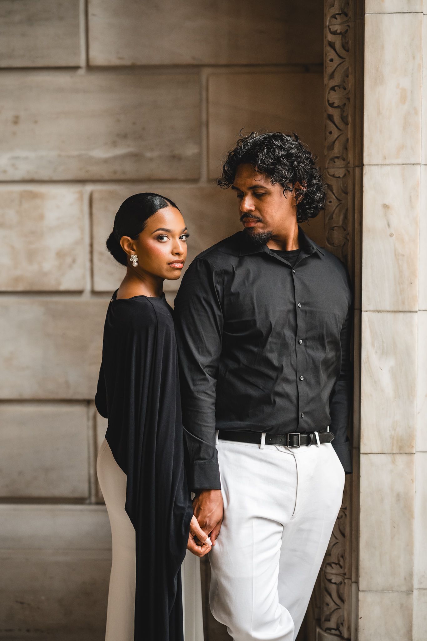 Elegant couple in black and white outfits posing against a stone wall, holding hands in a sophisticated urban setting.