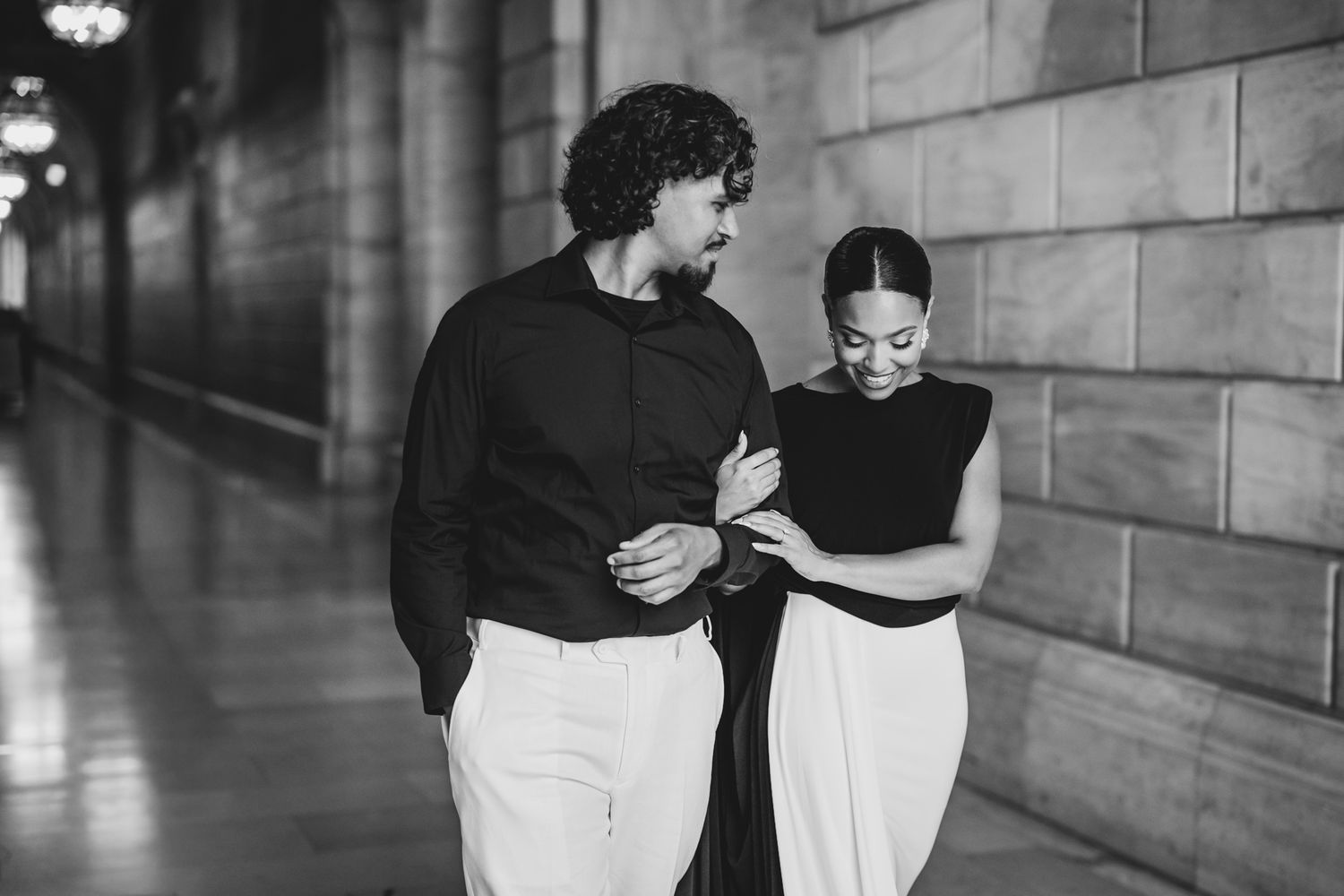 Black and white photo of an elegant couple walking together in a grand hallway, woman in white skirt and man in black shirt.