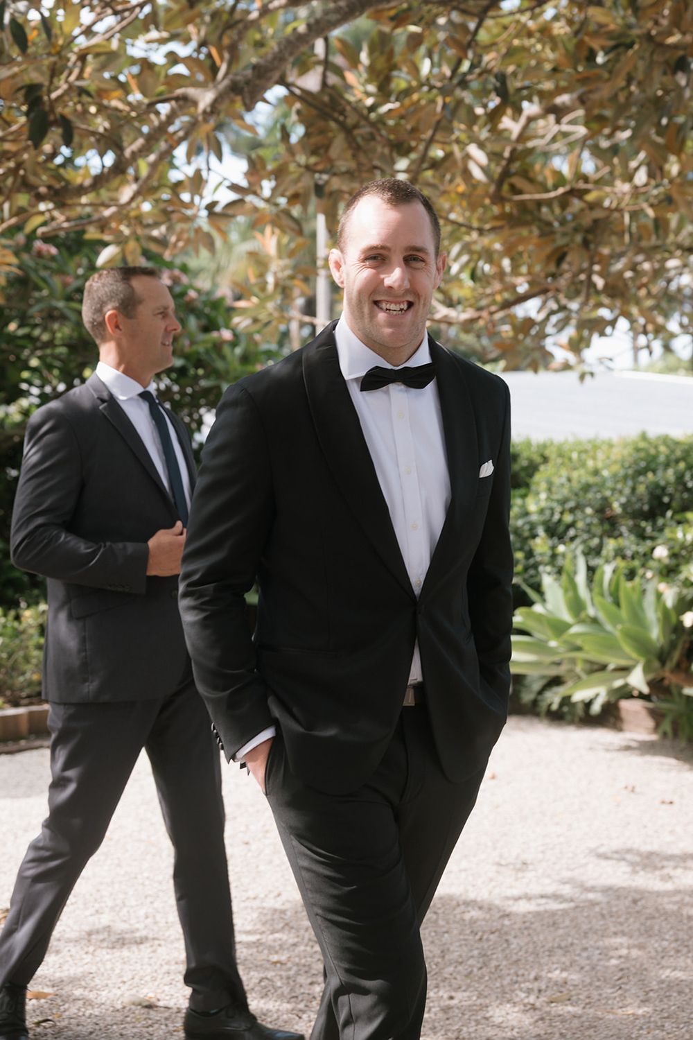 Smiling groom in black tuxedo and bow tie walks outdoors with groomsman in dark suit behind him.