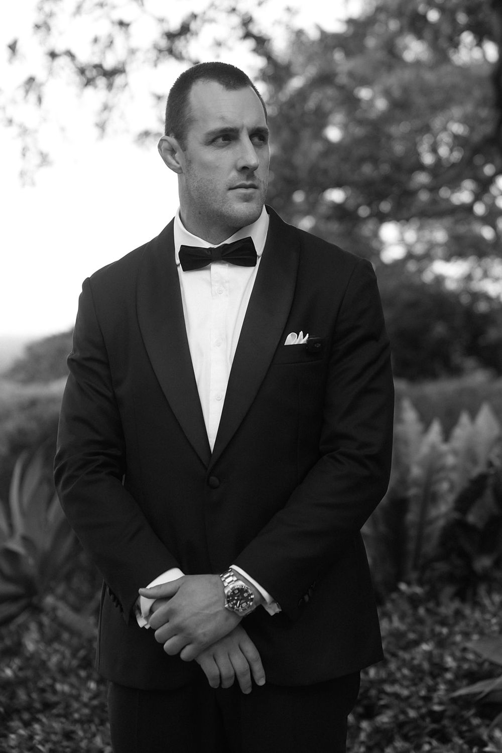 Black and white portrait of a groom in a tuxedo with bow tie and pocket square, standing outdoors.