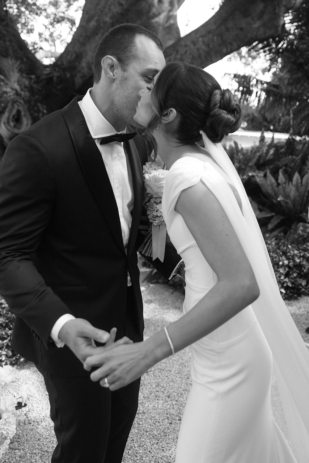 Black and white photo of a bride and groom sharing their first kiss while holding hands at an outdoor wedding ceremony.