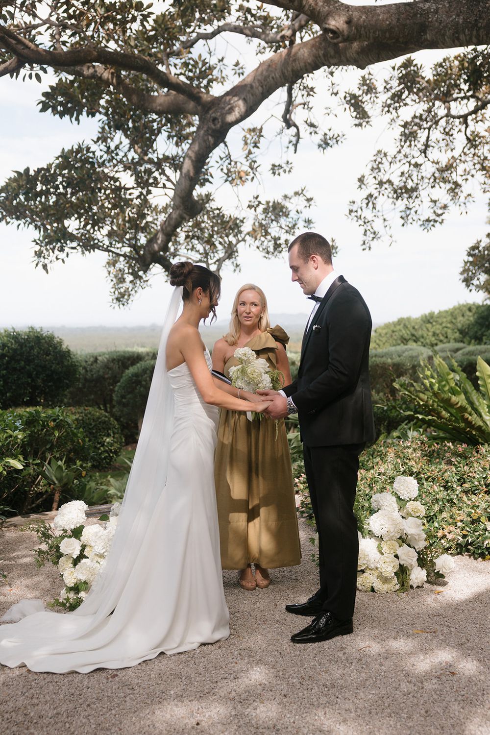 Bride and groom exchange vows outdoors under a tree, officiated by a woman in gold dress, with white floral arrangements.