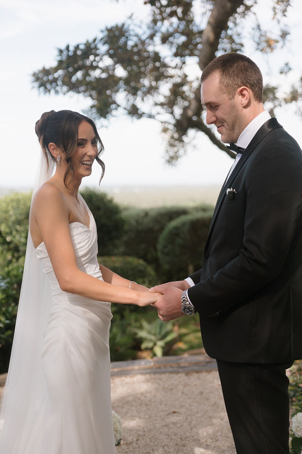 Bride and groom holding hands and smiling at each other during outdoor wedding ceremony in formal attire.