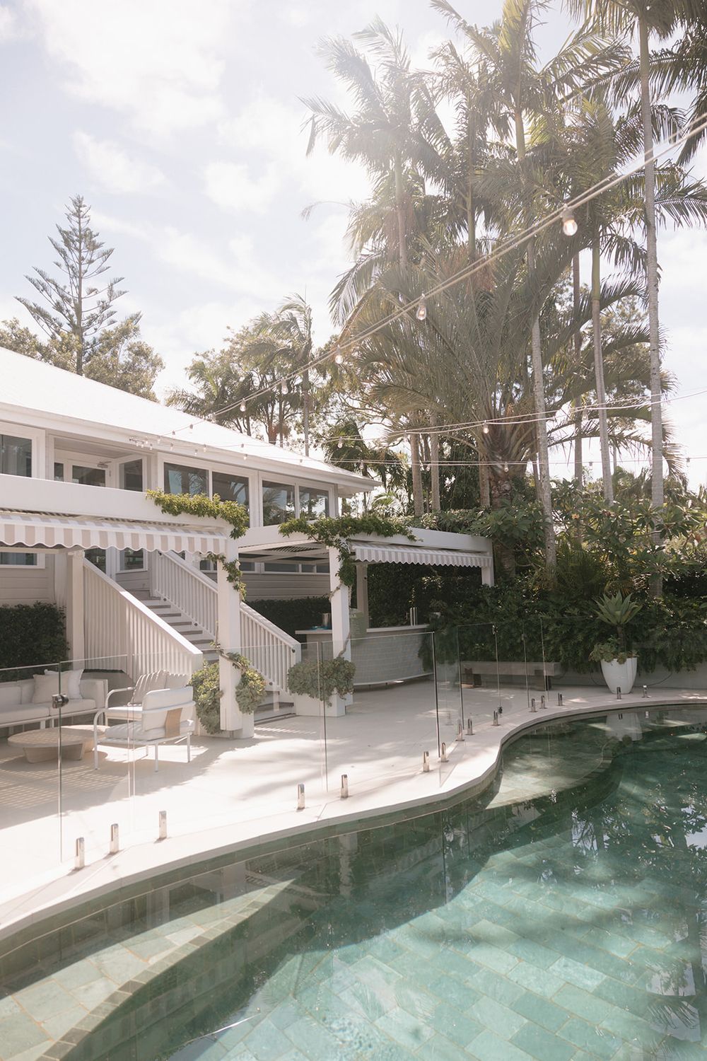 Elegant resort-style pool area with glass fencing, white building, striped awnings, and lush tropical palm trees.