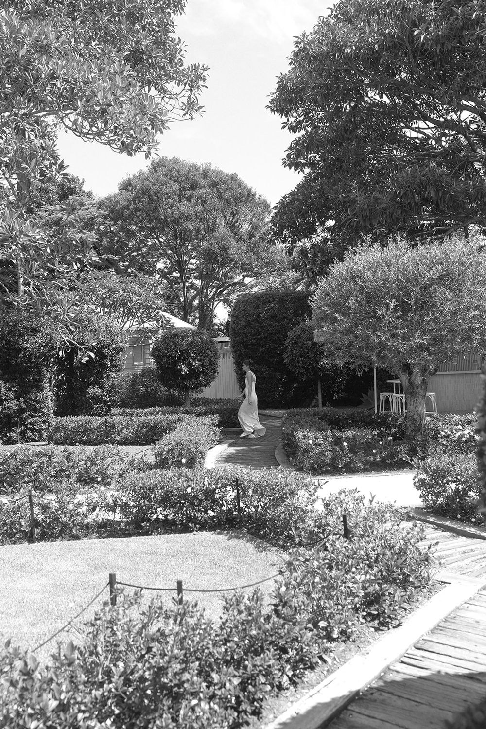 Black and white photo of a formal garden with manicured hedges, flowering plants, and a statue along a winding path.