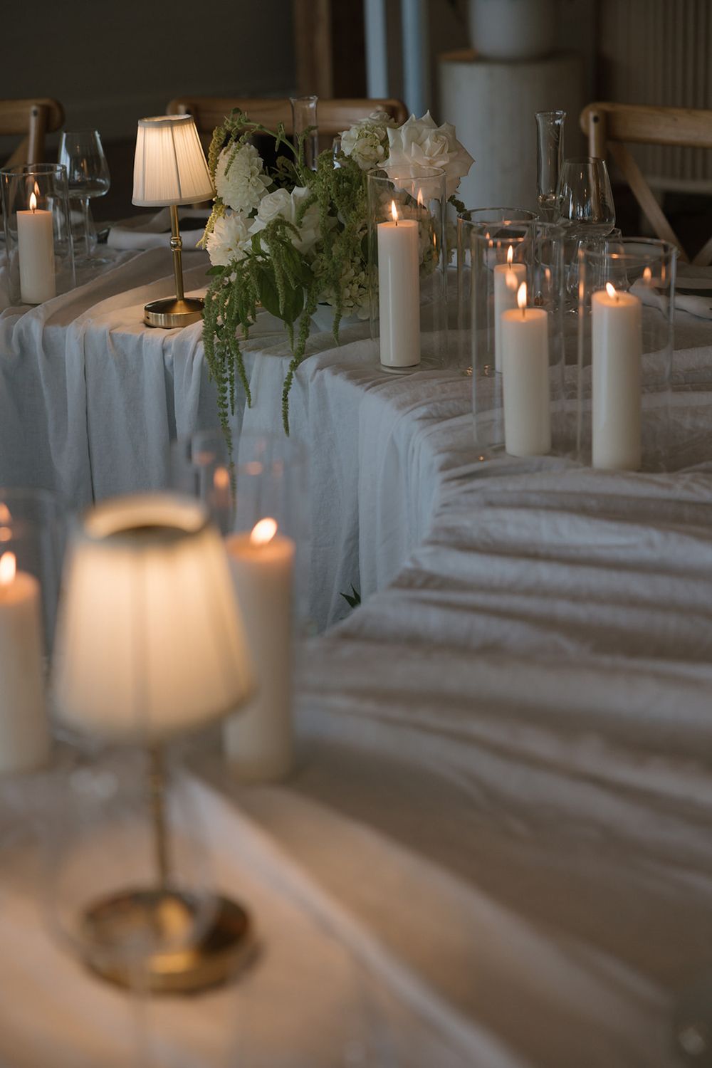 Elegant wedding reception table with white candles, floral centerpiece, and soft candlelight on a draped white tablecloth.