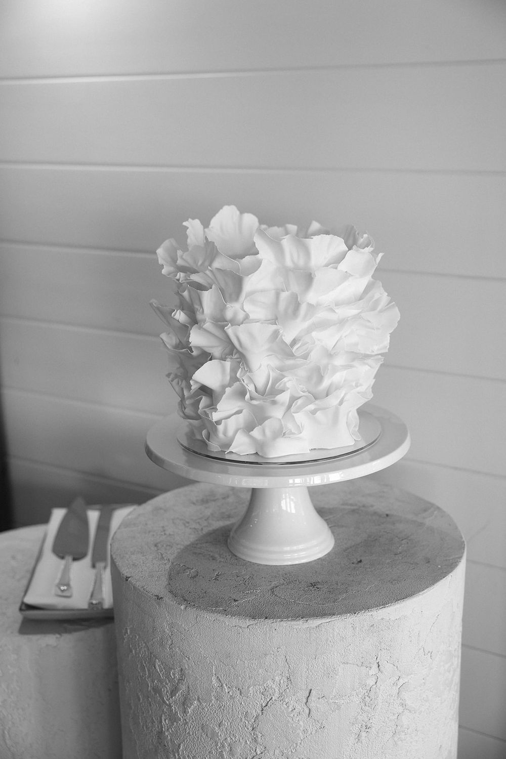 Elegant white ruffled wedding cake on a pedestal stand displayed on a concrete pillar in black and white photography.