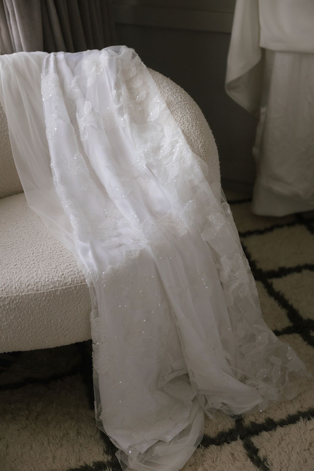 Delicate white lace wedding veil with pearl details draped over a white boucle armchair.
