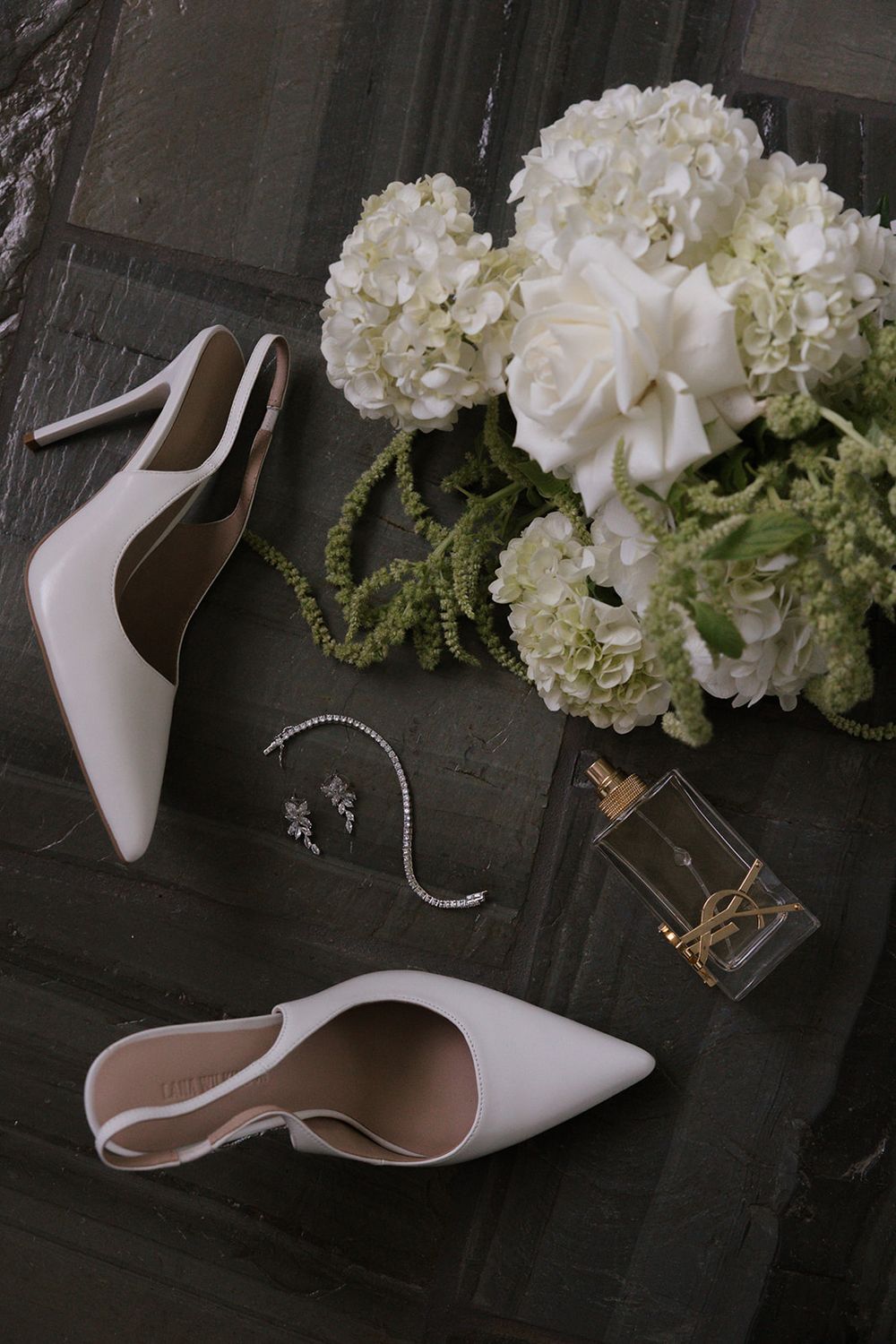 White bridal heels, diamond jewelry, YSL perfume, and white hydrangea bouquet arranged on dark surface.