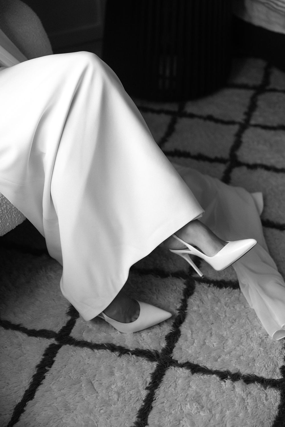 Black and white photo of a bride in a white wedding dress putting on elegant white heeled shoes on a patterned rug.