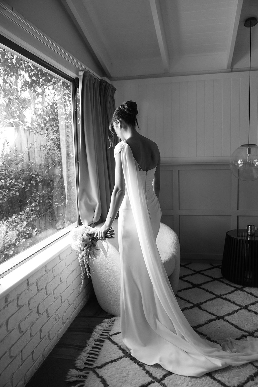 Elegant bride in a backless white gown with train gazing out a window in black and white.