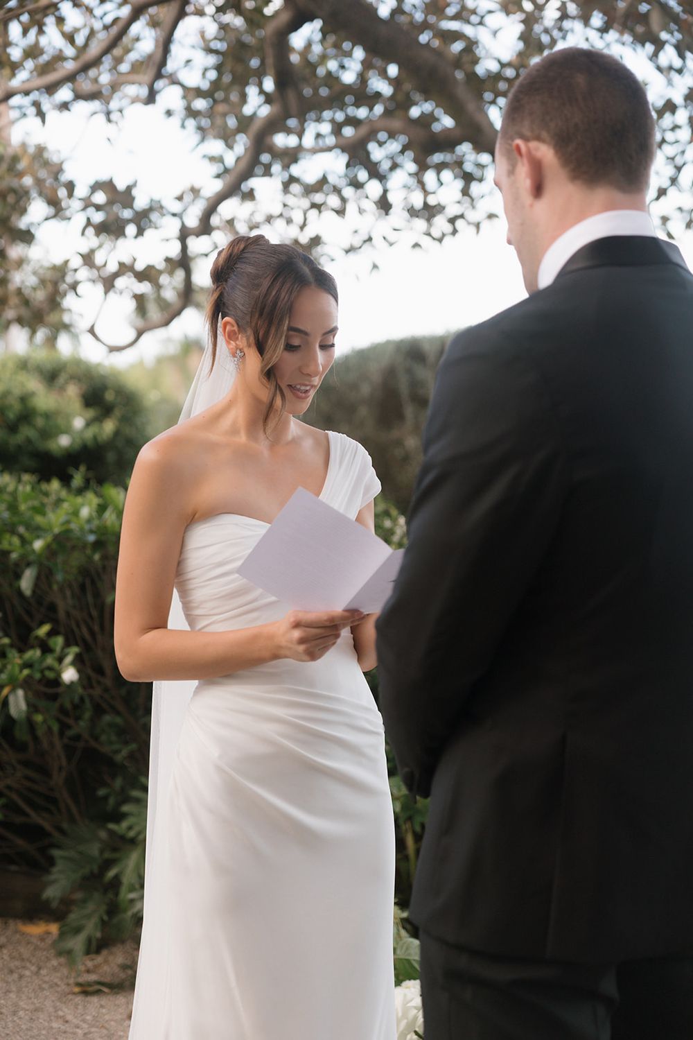 Bride in white one-shoulder gown reads vows from paper to groom in black suit during outdoor wedding ceremony.