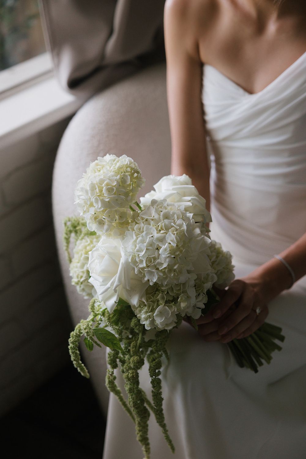 Bride in white dress holding elegant bouquet of white hydrangeas and roses with green amaranthus accents.