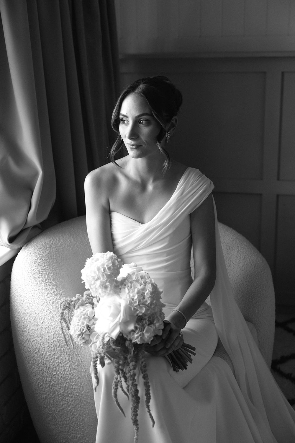 Black and white portrait of a bride in a white gown holding a bouquet, seated in a chair by a window.