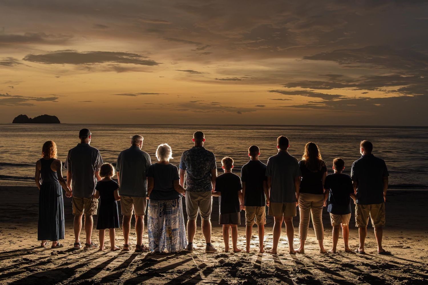 Large family standing together on a beach at sunset, silhouetted against a golden sky over the ocean.