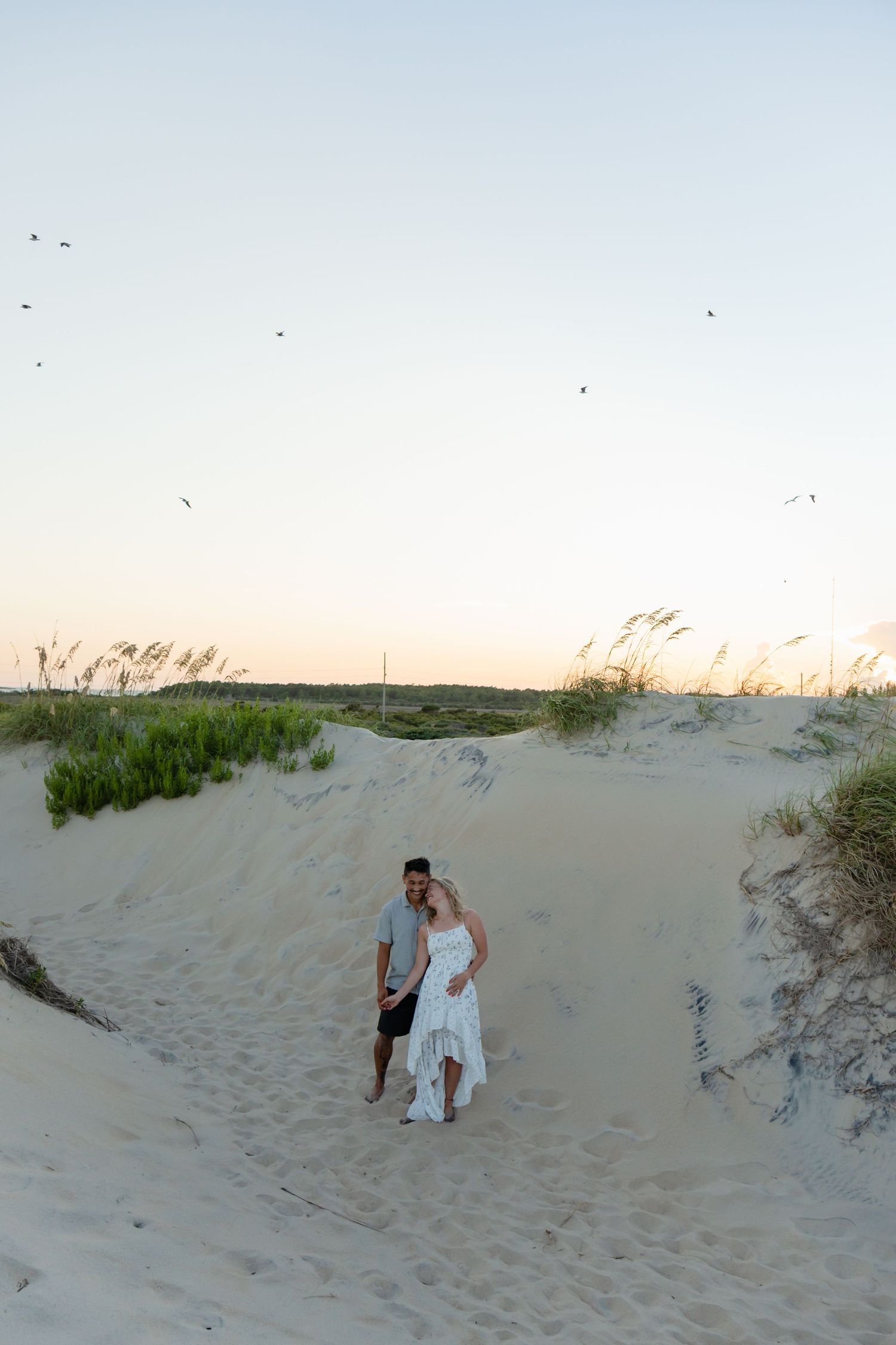 Couple sharing a romantic kiss on the beach, woman with tattoos in pink crop top embracing man in white shirt.