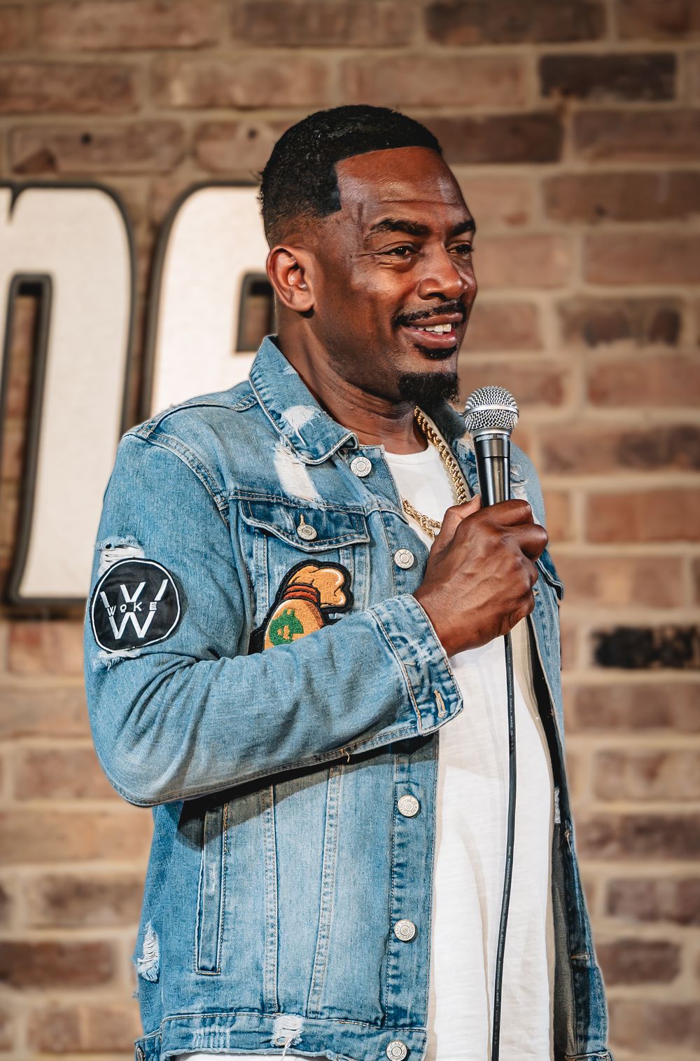 Bill Bellamy black comedian. Man in denim jacket holding microphone performing stand-up comedy on stage in front of brick wall.