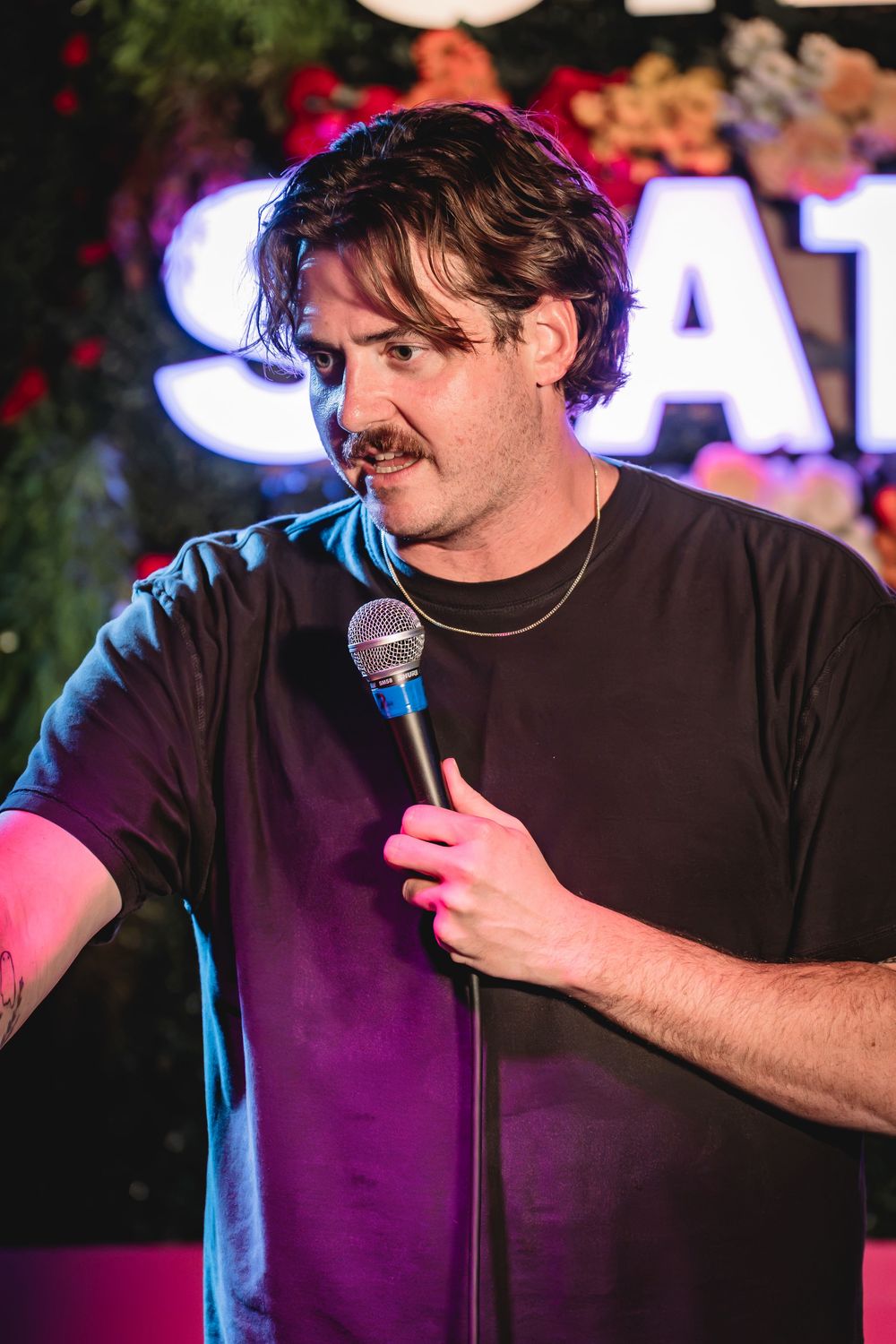 Dylan Carlino the comedian. Man with mustache performing stand-up comedy on stage, holding microphone under colorful neon lights.