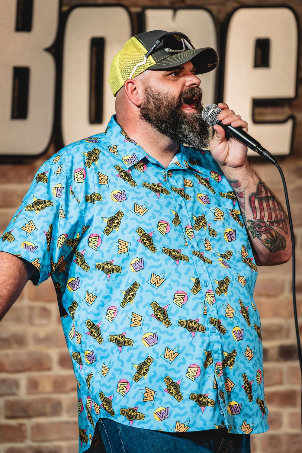 Graeson Mcgaha the comedian. Funny Bone Tampa. Bearded man in colorful blue novelty print shirt and cap speaks into microphone on stage.