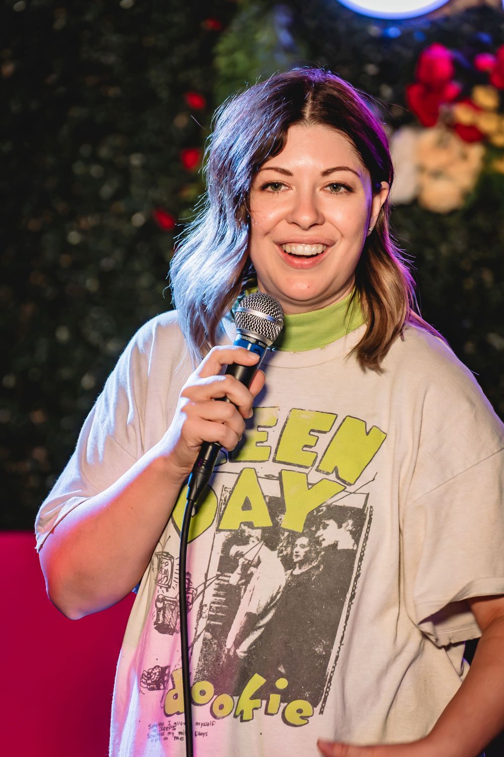Jessie "Jetski" Johnson. smiling woman holding microphone on stage wearing Green Day t-shirt performing stand-up comedy at colorful venue.