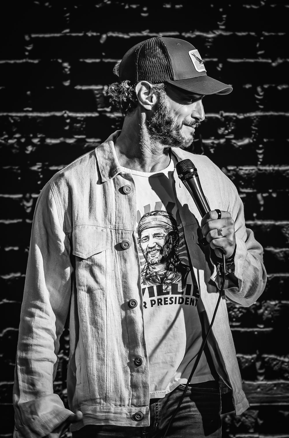 Josh Wolf comedian. Black and white photo of a bearded man in a trucker hat and denim jacket performing stand-up comedy with a microphone.