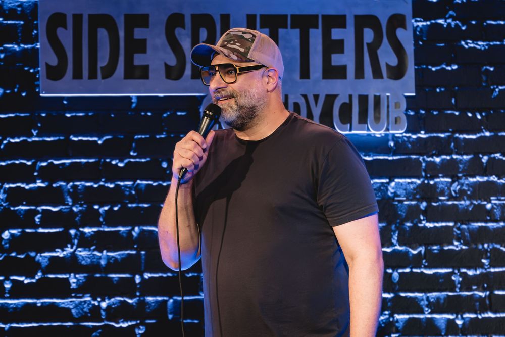 Kurt Metzger the Comedian performing stand-up at Side Splitters Comedy Club, holding microphone on stage with brick wall backdrop.