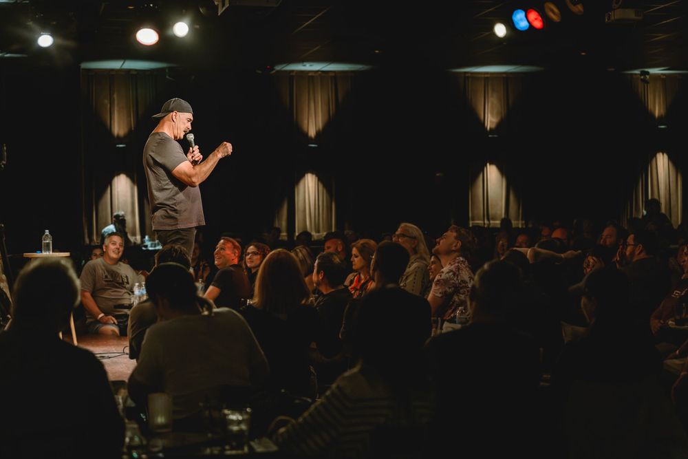 Comedian performing stand-up on stage before a large seated audience in a dimly lit comedy club.