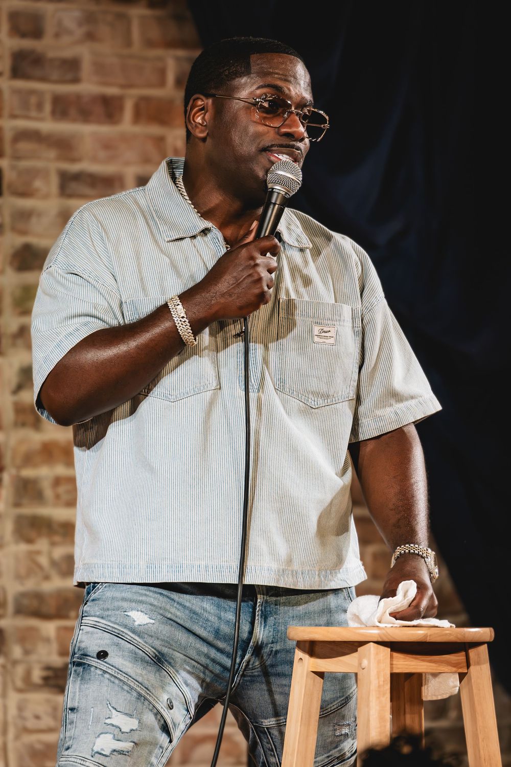 Skeet Carter the comedian. Man in gray shirt and jeans holds microphone on stage during stand-up comedy performance in front of brick wall.