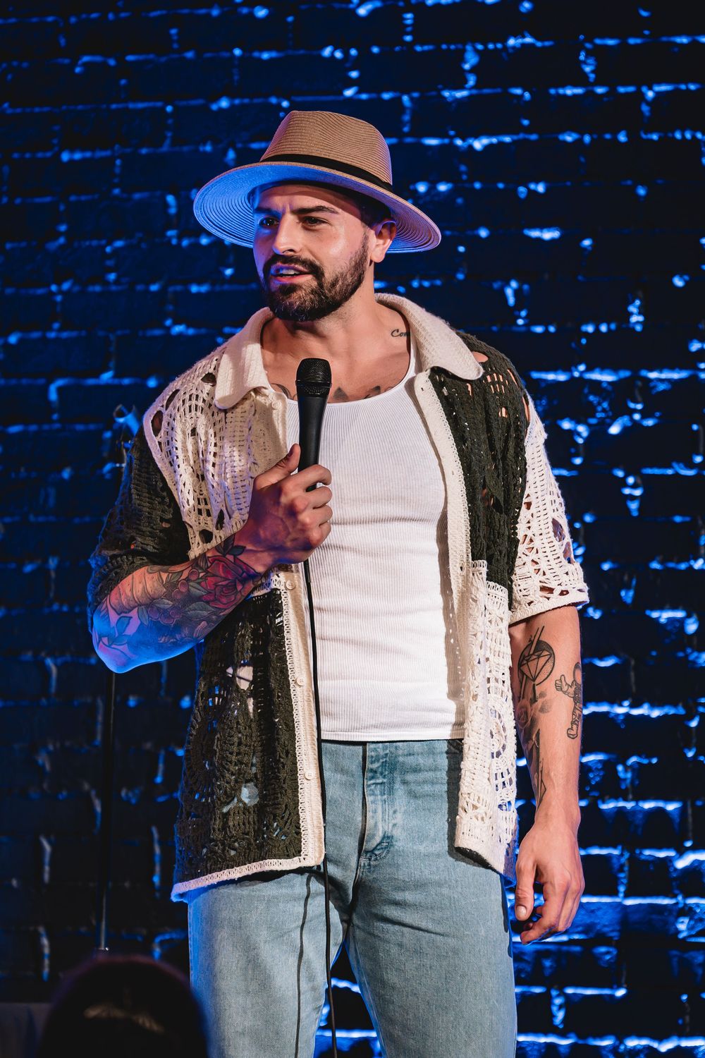 Zako comedian Tattooed male performer in a tan fedora, white tank top, and open crochet shirt holds a microphone on a blue-lit stage.