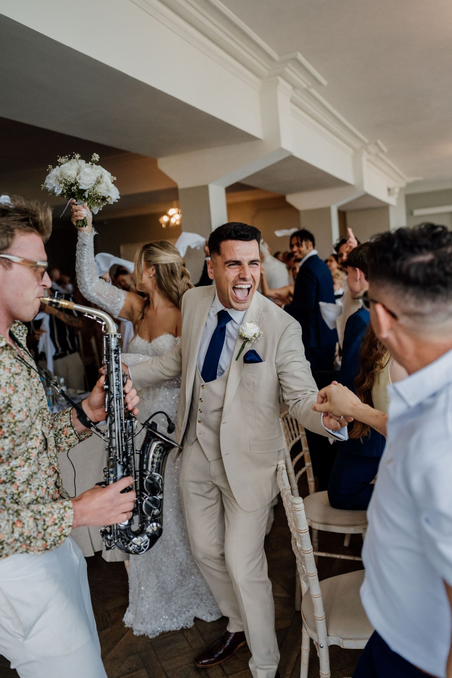 Groom in beige suit celebrating joyfully with saxophonist during lively wedding reception entrance.