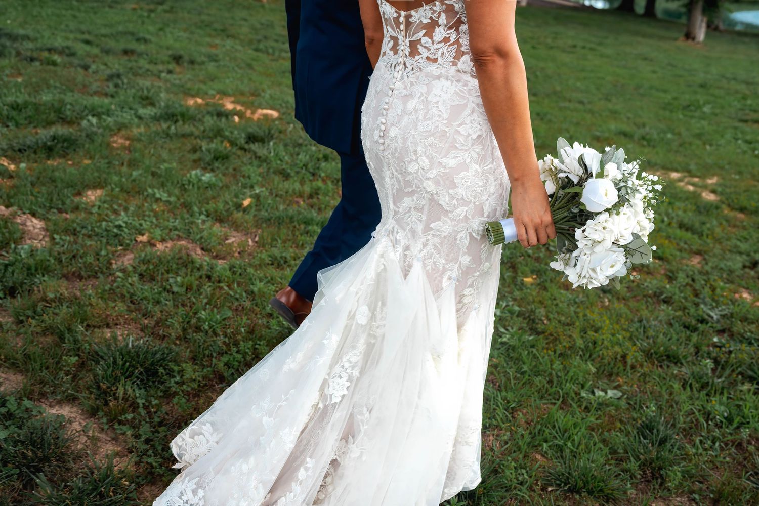 Bride in lace mermaid wedding gown holding white bouquet walks with groom in navy suit on green grass.