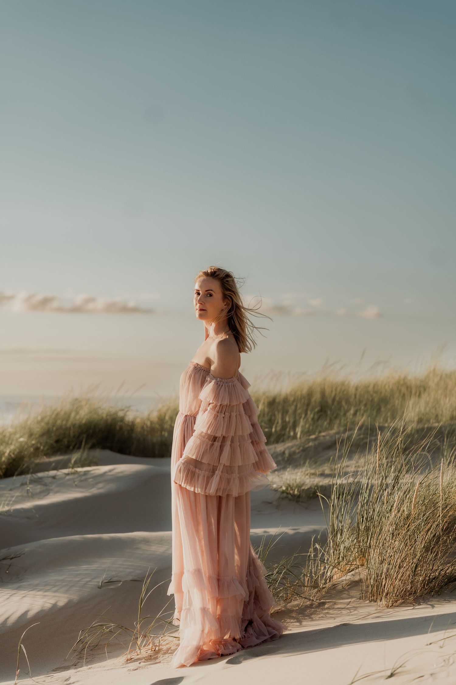 Woman in pink ruffled gown standing on sandy dunes with coastal grass and soft sky backdrop.