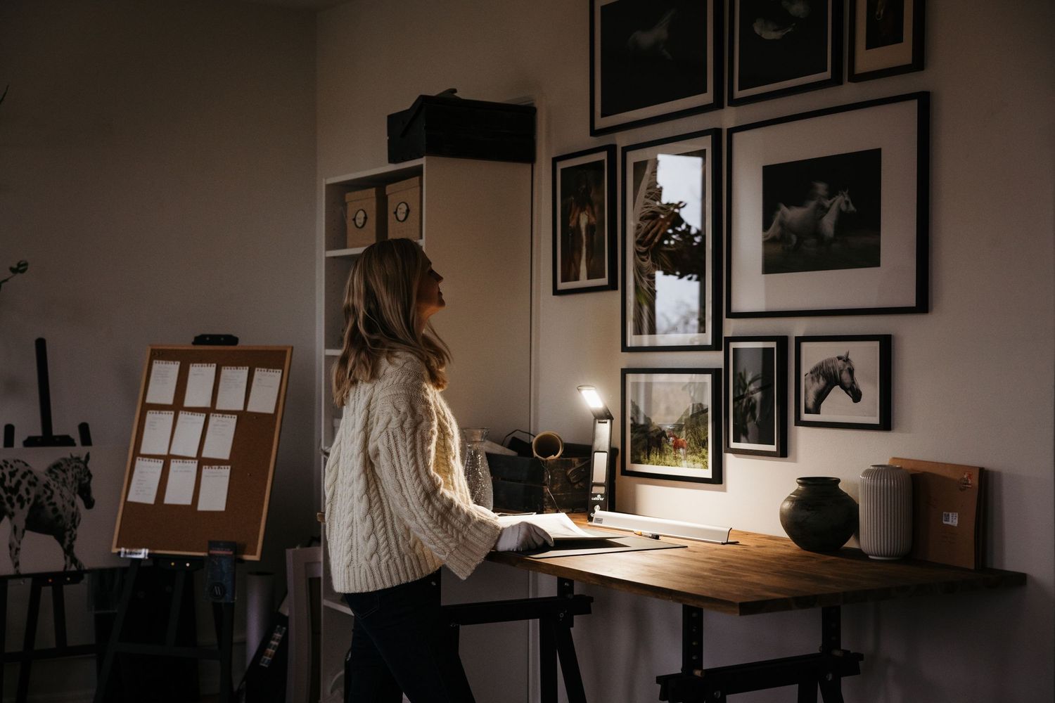 Woman in white sweater standing at wooden desk admiring framed art gallery wall in dimly lit home studio.