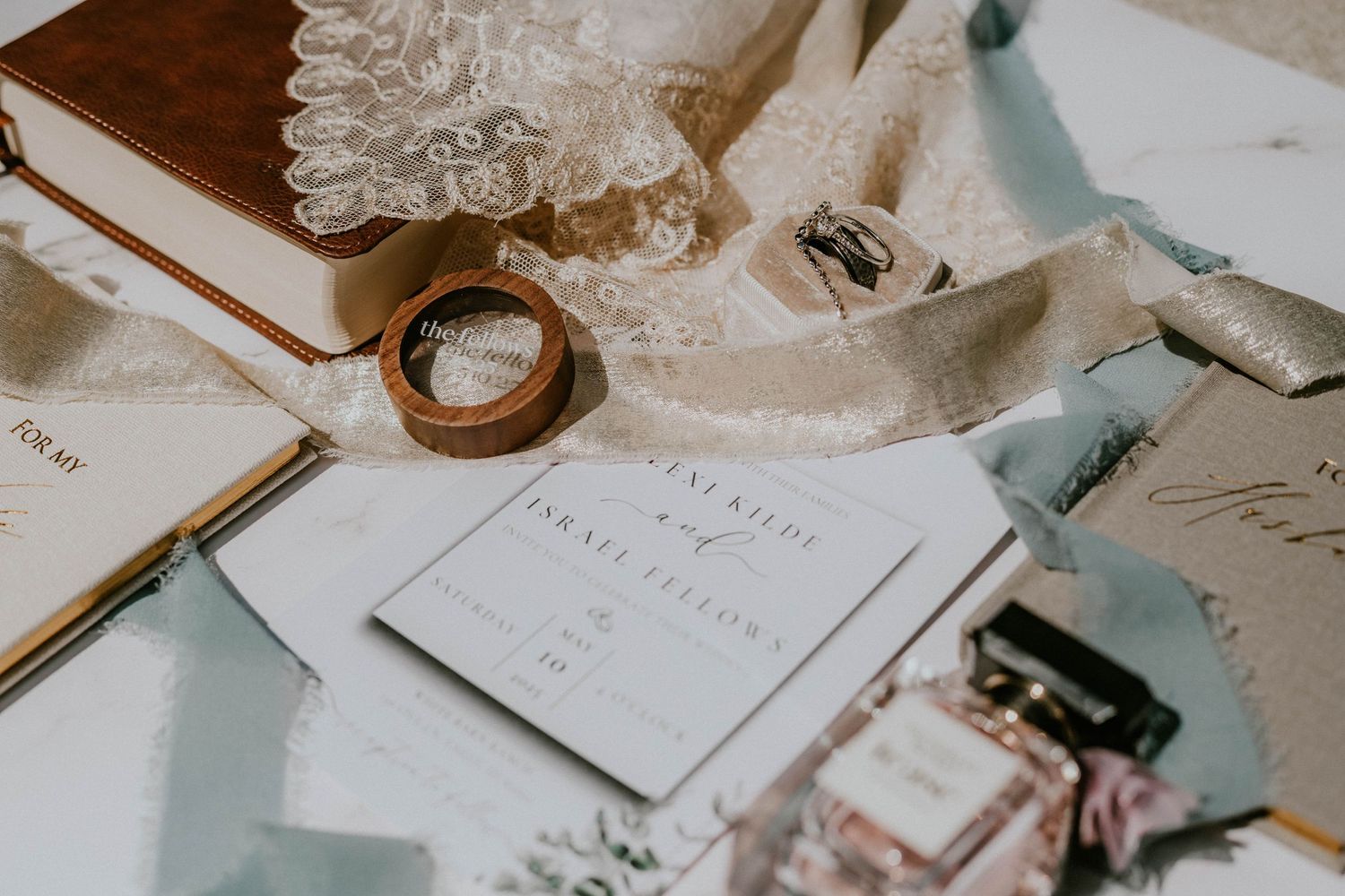 Elegant wedding flat lay with pearl necklace, gold ring box, ribbon, champagne glass, and wedding invitations on dark surface.