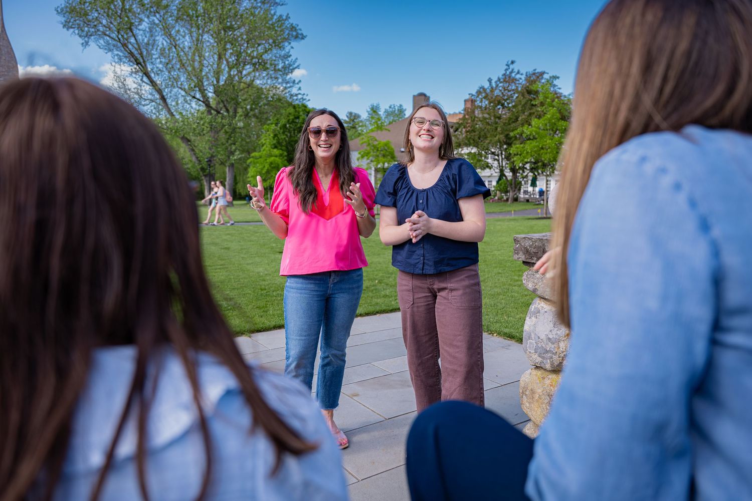 Two women smiling and talking outdoors on a sunny campus path surrounded by green trees and grass.