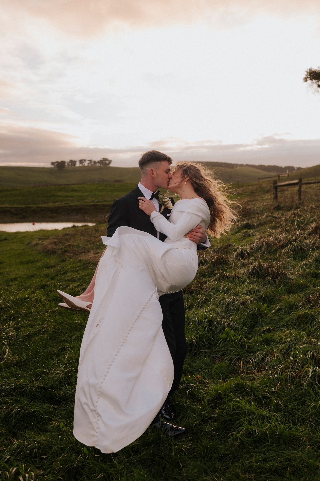 Groom lifts bride in white wedding dress for a romantic kiss at sunset on a lush green countryside hillside.